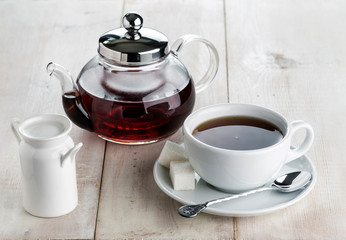 a glass kettle, tea cup of hot black tea and a milk jug on a wooden table