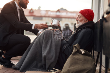 Kind man in suit hunkered down to homeless and give money donation, one dollar bill to beggar male