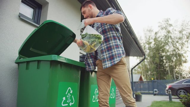 Caucasian Man is Walking Outside His House in Order to Take Out Two Plastic Bags of Trash. One Garbage Bag is Sorted into Biological Food Waste, Other is Thrown into Recyclable Bottles Garbage Bin.