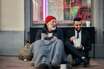 Smiling rich and poor men together sitting on street and eat while speaking. Happy despite social...