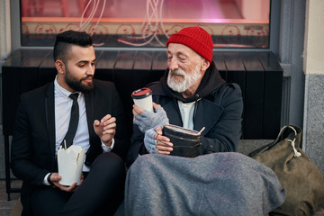 Young beardy man in suit sitting with beggar on floor on street and give cup of coffee. Different...
