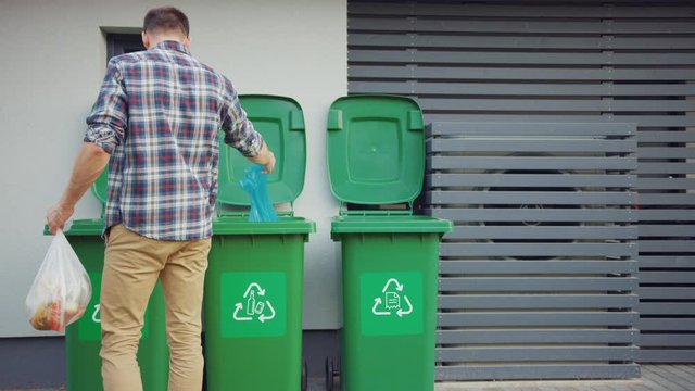 Caucasian Man is Walking Outside His House in Order to Take Out Two Plastic Bags of Trash. One Garbage Bag is Sorted into Biological Food Waste, Other is Thrown into Recyclable Bottles Garbage Bin.