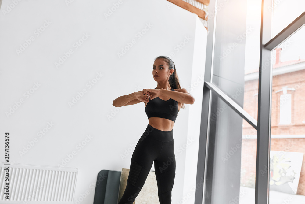 Wall mural Slim girl doing exercises standing at gym. Dressed in black clothes. Background window