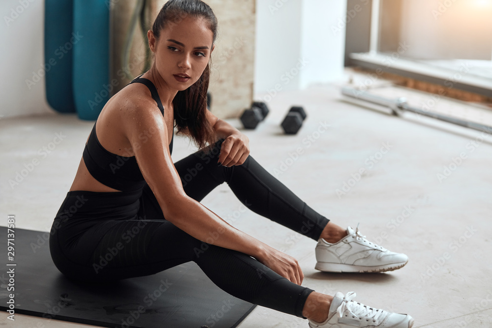 Wall mural Beautiful caucasian girl in sportswear sit tired, have rest after exercising in gym. Window background