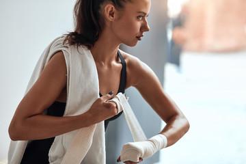 Side view on fit female tying bandages on her hands before fitness training, towel on shoulders. Window background
