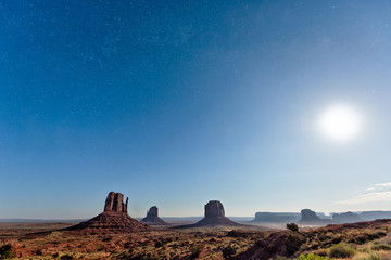 Obraz na płótnie Canvas Famous buttes wide angle view in Monument Valley during blue twilight dark night with stars in Arizona dust road and bright moon in sky