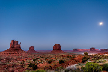 Famous mesa butte formations with red rock color in Monument Valley canyons during twilight dark night in Arizona with cars on dirt road and moon in sky
