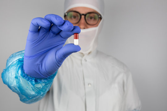 Young woman with glasses wearing white sterile protective clothing and blue gloves  holding a red and white pill in front of her