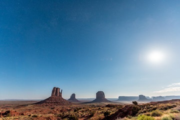 Fototapeta premium Famous wide angle view in Monument Valley of butte during blue twilight dark night with stars in Arizona dust road and bright moon in sky