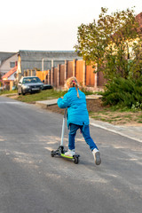 back view. girl in a blue jacket riding a scooter. blond hair color. she rolls her foot on the board.