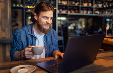 Young bearded freelancer checking email via laptop while having morning coffee in a loft bar