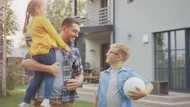 Portrait of a Happy Family of Three: Father, Daughter, Son. They Are Posing In Front of Camera on a Lawn Next to Their Country House. Dad is Holding the Girl in His Arms. Boy is Holding a Football.
