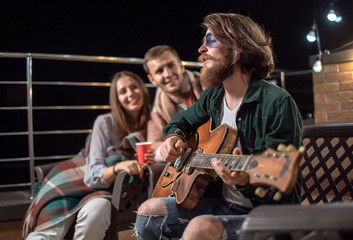 Young couple listening to their friend's romantic guitar-accompanied song on a cozy balcony after sunset
