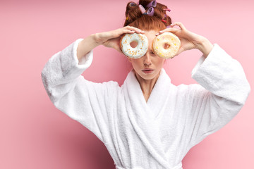 Cute redhaired woman holding sweet donut, happy after shower in bathrobe and curlers on hair, closed both eyes. Isolated over pink background