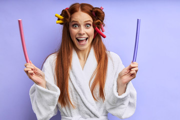 Young caucasian woman in bathrobe with curlers on hair posing, look at camera. Portrait, isolated over purple background