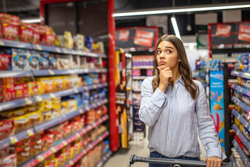 Young woman looking confused choosing products from an aisle at the supermarket walking with her...