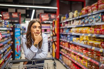 Young woman staying confused in grocery store. Confused woman doesnt know what to buy. Young woman...