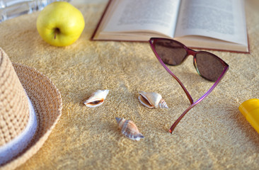 Straw hat, seashells, sunburn cream, sunglasses, apple, bottle with water and opened book on beige beach towel. Soft evening sunlight.