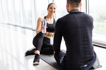 Tired fitness couple having rest and talking, drinking water at gym. Man and woman sitting on floor, break after training,