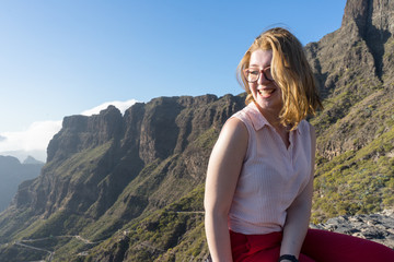 Naklejka premium Serene Cute Tourist woman, laughing and admiring the freedom and the landscape in the mountain, sitting in a rock. Model sit and smiling in the nature with excitement