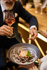 Close image of hand with glass of champagne, sweet dessert on grey dish. Beared man in tuxedo drinking