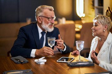 Senior couple spend time in restaurant, speaking, use modern technologies - laptop, tablet, IQOS. Elegant grey-haired man in tuxedo, woman in white blazer