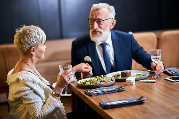 Modern senior couple in love, look at each other, speak. Stylish people in tuxedo and blazer use modern technologies, devices, iqos, electronic cigarette. Man takes a woman's hand