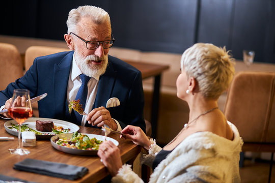 Pretty Lady And Elderly Grey Haired Man In Tuxedo Having Friendly Conversation In Restaurant. Colleagues After Work Discuss And Eat