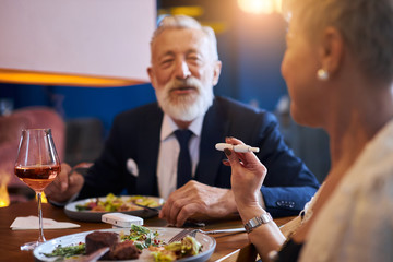 Senior caucasian bearded male sits and look at woman with e-cigarette, iqos in restaurant hotel. Elegant man in tuxedo. Modern technologies concept