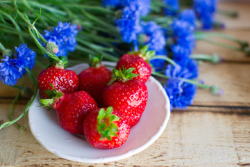 Seasonal summer flowers (blue cornflowers) and fruits (strawberries) on rustic scorched wooden table