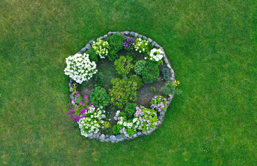 Aerial drone top down view on stylish round flowerbed surrounded by rock wall with violet, white and yellow blooming flowers. Grass as background