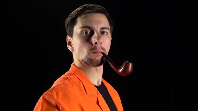 Close up of man in a bright suit stands and smokes with a pipe gazing intently at the camera