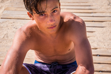 close up of teenager and caucasian boy sitted on the sand and on the wood at the beach - attractive and handsome young man looking at the camera in swimwear