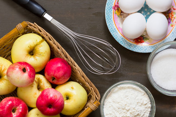 Top view of apples, eggs, sugar, flour and whisk on wooden background