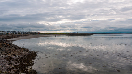 View of Salthill beach and Mutton island on a cloudy day, with rocks in the foreground and mountains in the background.