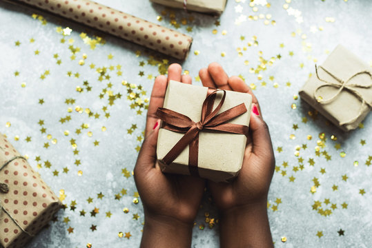 A dark-skinned girl gives a gift made with her own hands. Hands holding a craft gift with a brown ribbon on a gray background with a few small gifts. Flat lay