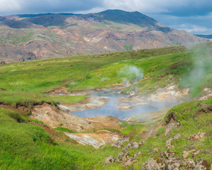 Reykjadalur valley with hot springs river and pool with lush green grass meadow and hills with geothermal steam. South Iceland near Hveragerdi city. Summer sunny morning, blue sky.