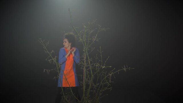 Crazy girl near branches at dark studio with haze looks around with scared, sees something straight and leaves with smiling