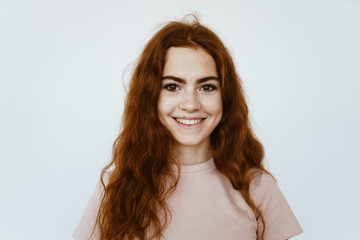 Red-haired girl with an open smile and brown eyes. a flesh-colored t-shirt. dimples on the cheeks, hair curly. studio background light gray - 297165340