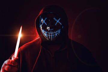 Man with knife in hand and angry and scary lighting neon glow mask in hood on dark red background....