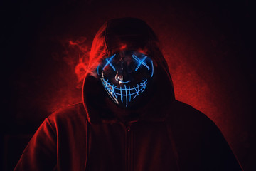 Man in angry and scary lighting neon glow mask in hood on dark red background. Halloween and horror...