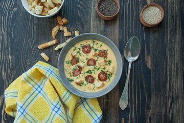 Cheese soup with grilled sausages and herbs. Cream soup served in a plate. Dark wooden background.