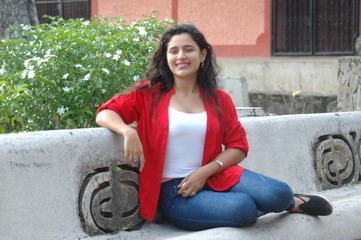Young black hair wearing a red shirt and blue trousers facing the camera smilingly sitting on a seminal bench with a small tree in the background.
