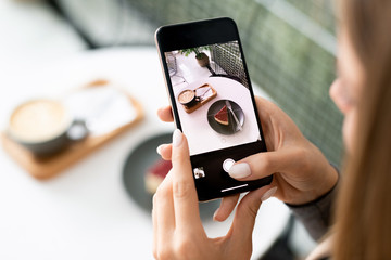 Young woman with smartphone taking photo of cappuccino and dessert