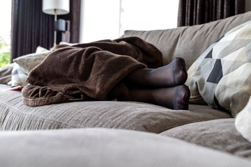 A potrait of a girls black pantyhose feet while she is sleeping in the couch in the living room under a brown cosy blanket.