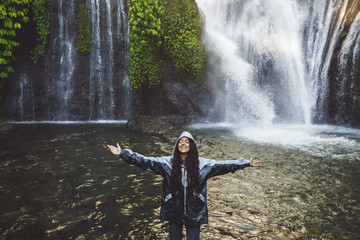 Young happy woman enjoying waterfall in Bali. Wearing grey raincoat from water spray. Travel in Indonesia.