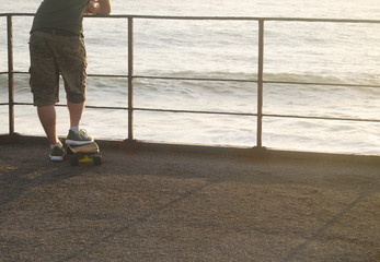 Pessoa com um skate parada a ver o mar ao fim do dia - obrazy, fototapety, plakaty