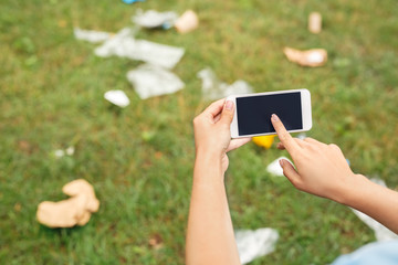 Leisure Outdoors. Young girl sitting in park taking photo of wastes on smartphone touching screen close-up blurred