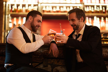 Side view portrait of two businessmen drinking whiskey and clinking glasses while sitting by bar counter and relaxing after work