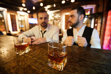 Portrait of two ruffled businessmen drinking in bar after work, focus on two whiskey glasses on counter, copy space